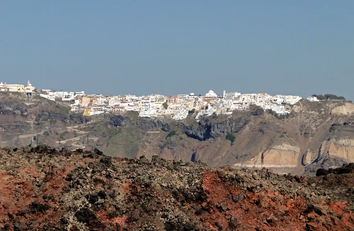 Fira on the caldera rim seen across red lava rock on Nea Kameni during a Santorini catamaran cruise