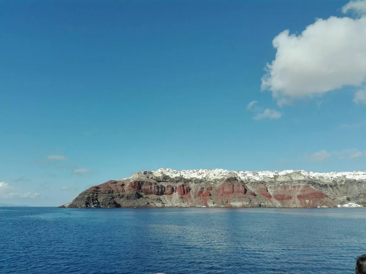 Oia clinging to the red and black caldera cliff seen from the water on a Santorini catamaran cruise